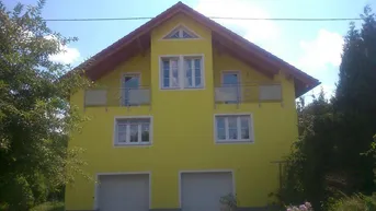Gelbes Einfamilienhaus mit Satteldach, zwei Garagen und kleinen Balkonen, umgeben von viel Grün.