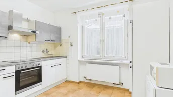 Helle Küche mit weißen Schränken, Fliesenboden und Fenster mit Vorhängen.
