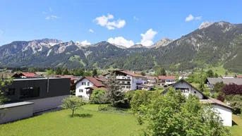 Weitläufiger Blick über die grüne Landschaft und das Dorf mit majestätischen Bergen im Hintergrund.