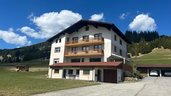 Mehrstöckiges Gebäude mit Holz-Balkonen, umgeben von grünen Wiesen und Bergen unter blauem Himmel.