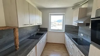 Moderne Küche mit hellen Holzschränken, dunklen Arbeitsplatten und einem Fenster mit Bergblick.
