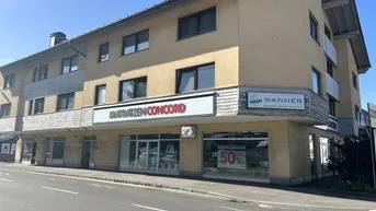 Gebäudeansicht mit Ladenlokalen im Erdgeschoss und Wohnungen darüber an einer Straße gelegen.