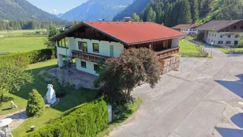 Traditionelles Haus mit rotem Dach und Holzbalkonen, umgeben von grüner Landschaft und Bergen.