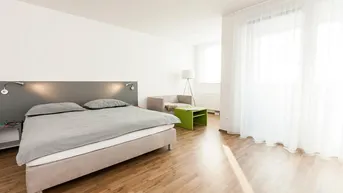 Helles Schlafzimmer mit Holzboden, großem Bett und einem kleinen Sitzbereich am Fenster.