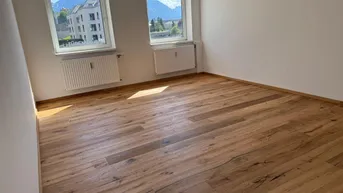 Heller, leerer Raum mit Holzdielenboden, zwei Fenstern und Bergblick.