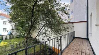 Langer Balkon mit Holzboden und schwarzem Geländer, bietet Blick auf den grünen Innenhof.