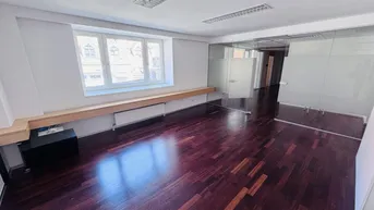Geräumiges Büro mit dunklem Parkettboden, weißen Wänden und modernen Glasabtrennungen, hell durch Fenster.