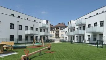 Moderne Wohnanlage mit weißer Fassade und einem gemeinschaftlichen Gartenbereich inklusive Spielplatz für Kinder.
