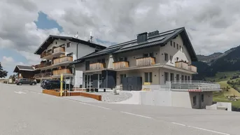 Modernes Gebäudeensemble mit traditionellen Holzbalkonen und Photovoltaikanlage in alpiner Umgebung, umgeben von grünen Hügeln.