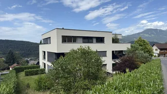 Modernes Mehrfamilienhaus mit Flachdach und großen Fenstern in grüner Umgebung mit Bergblick.