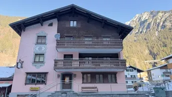 Traditionelles Gebäude mit rosa Fassade, Holzbalkonen und Satteldach, umgeben von schneebedeckten Bergen.
