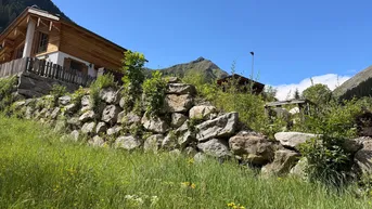 Holzhaus im Chalet-Stil mit Steinmauer und Bergblick unter blauem Himmel.