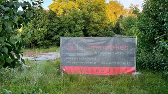Grundstück mit Verkaufsplakat, umgeben von üppigem Grün und Bäumen unter blauem Himmel.
