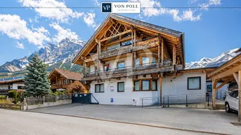 Traditionelles Gebäude mit Holzfassade und Bergblick, umgeben von einer gepflasterten Fläche.