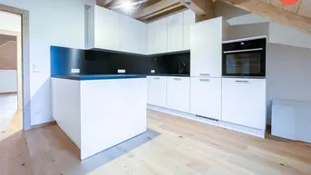Moderne Küche mit weißen Schränken, dunkler Arbeitsplatte und sichtbaren Holzbalken unter dem Dach.