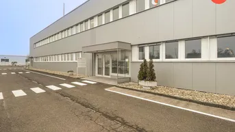 Eingangsbereich eines modernen Gewerbegebäudes mit Glasfassade, Zufahrtsstraße und Parkplätzen im Hintergrund.