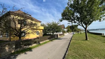 Straße entlang eines gelben Hauses mit Blick auf den Fluss und die gegenüberliegende Landschaft.
