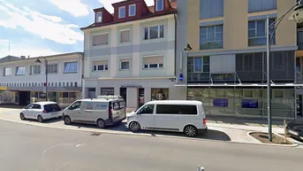 Ansicht des Mehrfamilienhauses von der Straße mit geparkten Fahrzeugen und Geschäften im Erdgeschoss.