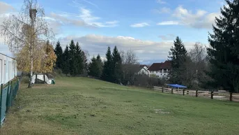 Weitläufiges grünes Grundstück mit Bäumen und einem Haus im Hintergrund unter bewölktem Himmel.