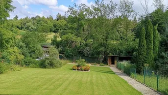 Gepflegter Rasenbereich mit Blumenbeet und Gartenhaus, umgeben von üppiger Vegetation und Bäumen.