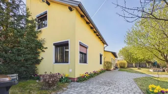 Gelbes Einfamilienhaus mit gepflasterter Auffahrt und grüner Bepflanzung im Vorgarten.