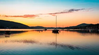 Malerischer Sonnenuntergang über einem ruhigen See mit Segelbooten und Bergen im Hintergrund.