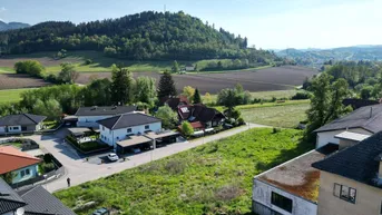 Luftaufnahme einer ländlichen Wohngegend mit Häusern, Feldern und bewaldeten Hügeln im Hintergrund.