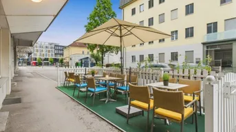 Außenbereich mit Tischen, Stühlen und Sonnenschirm, umgeben von einem weißen Zaun.