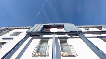 Moderne Fassade eines Wohngebäudes mit kleinen Balkonen und markantem Dach unter blauem Himmel.