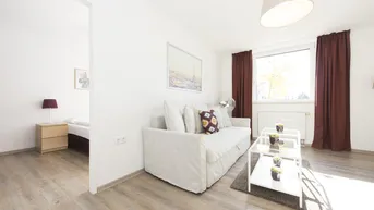Helles Wohnzimmer mit modernem Sofa, Holzboden und Blick in ein angrenzendes Schlafzimmer.