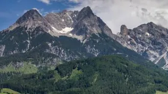 Majestätische Berge mit dichten grünen Wäldern und schneebedeckten Gipfeln unter einem bewölkten Himmel.