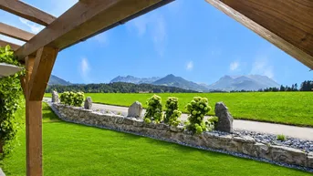Blick auf die Berge über eine grüne Wiese mit einer Holzpergola im Vordergrund.