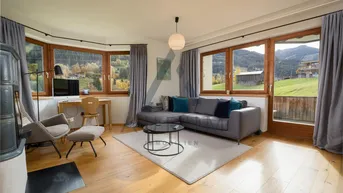 Helles Wohnzimmer mit gemütlichem Sofa, Schreibtischbereich und traditionellem Kachelofen, umgeben von großen Fenstern mit Bergblick.
