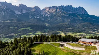 Panoramablick auf ein Gebäudeensemble in den Bergen, umgeben von grünen Hügeln und Wäldern.
