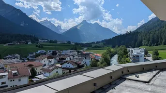 Weitläufiger Blick über ein grünes Tal mit Häusern, einem Fluss und majestätischen Bergen im Hintergrund.