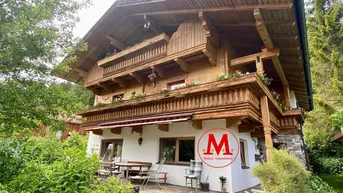 Traditionelles Holzhaus im alpinen Stil mit Terrasse und Gartenbereich, umgeben von üppigem Grün.
