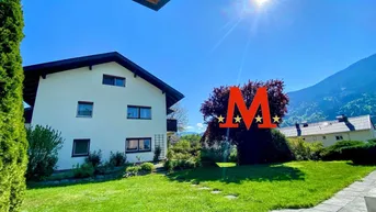 Weißes Einfamilienhaus mit großem Garten und Bergblick unter strahlend blauem Himmel.