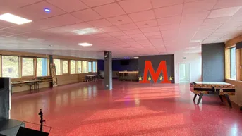 Großer Mehrzweckraum mit rotem Boden, vielen Fenstern und einer Bar im Hintergrund, ideal für Veranstaltungen.