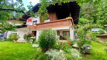Charmantes Haus mit gepflegtem Vorgarten und blühenden Pflanzen, umgeben von grüner Natur.