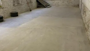 Großer, leerer Raum mit Betonboden, unverputzten Wänden und einer Holztreppe, die in den oberen Bereich führt.
