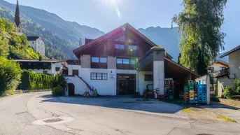 Alpines Gebäude mit Holzelementen und weißer Fassade, umgeben von Bergen und einem Kirchturm.