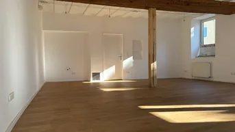 Geräumiger Innenraum mit Holzbalken, Holzboden und viel natürlichem Licht durch die Fenster.