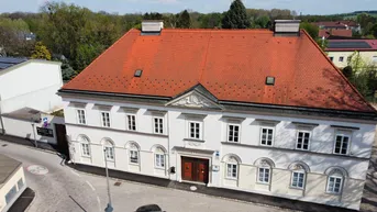 Historisches Gebäude mit weißer Fassade, rotem Ziegeldach und vielen Fenstern in einer grünen Umgebung.