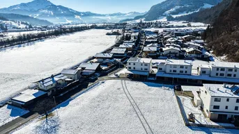 Luftaufnahme eines verschneiten Dorfes mit umliegenden Feldern und majestätischen Bergen im Hintergrund unter blauem Himmel.