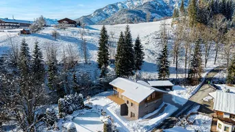 Luftaufnahme eines modernen Hauses in verschneiter Berglandschaft mit umliegenden Bäumen und Wegen.