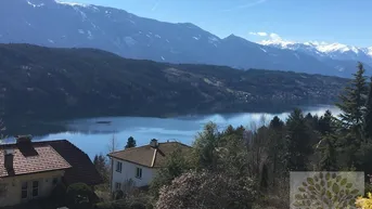 Panoramablick auf den See und die umliegenden Berge mit Häusern im Vordergrund unter blauem Himmel.