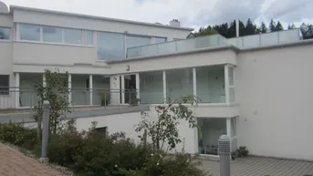 Modernes Gebäude mit großen Fenstern, Balkonen und gepflegter Außenanlage unter bewölktem Himmel.
