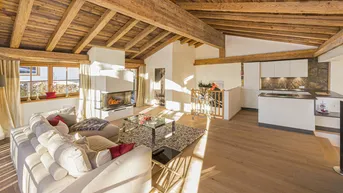 Lichtdurchfluteter Wohnbereich mit modernem Kamin, Holzbalkendecke und offener Küche im Hintergrund.