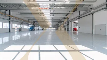 Große, helle Lagerhalle mit glänzendem Boden, Oberlichtern und Kranbahn für industrielle Nutzung.