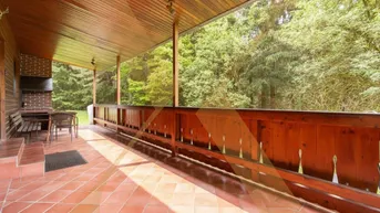 Geräumige überdachte Terrasse mit Fliesenboden, Holzbalken und integriertem Grillbereich, umgeben von viel Grün.